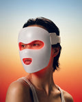 A woman wearing Morphace LED light therapy face mask that glows red-orange, shown against a warm orange-to-blue gradient background.